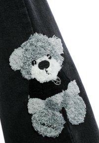 Parche de osito de peluche gris y blanco que lleva una camiseta negra, cosido sobre tela de mezclilla negra.