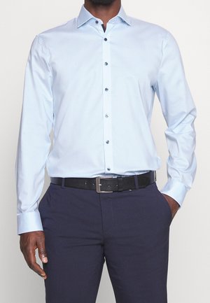 Chemise boutonnière bleu clair à manches longues, boutons foncés contrastants, associée à un pantalon foncé et une ceinture noire. Texture lisse.