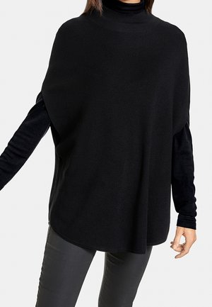 Kvinde iført en løs sort strikket ærmeløs poncho over en tætsiddende sort langærmet top og mørkegrå bukser, stående mod en hvid baggrund.