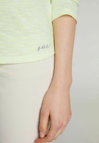 Detailní záběr na ruku a paži osoby vedle světle žlutého a bílého proužkovaného trička a béžových kalhot.