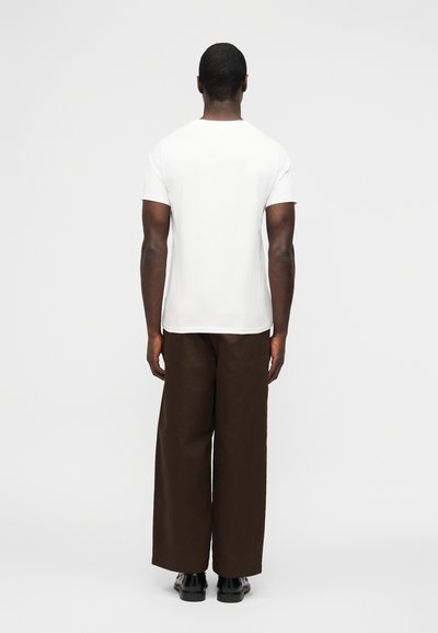 Άνδρας που στέκεται γυρισμένος ανάποδα, φοράει απλό λευκό κοντομάνικο πουκάμισο, φαρδιά καφέ παντελόνια και μαύρα παπούτσια, σε απλό φόντο.