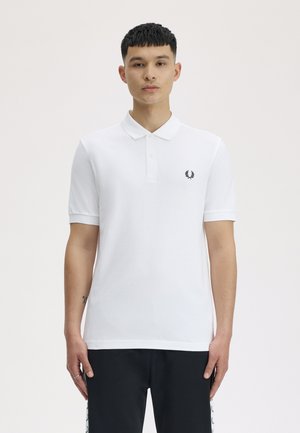 Jonge man die een wit poloshirt draagt met een klein zwart laurel-logo op de borst en een zwarte broek, staand tegen een effen achtergrond.