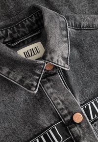 Kołnierz ciemnoszarej kurtki jeansowej z metalowymi guzikami oraz widoczną metką materiałową i logo z napisem "BIZUU" na kołnierzu i piersi.