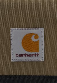 Firkantet hvidt stofmærke syet på groft brunt stof med orange Carhartt-logo og maroon Carhartt-tekst nedenunder.