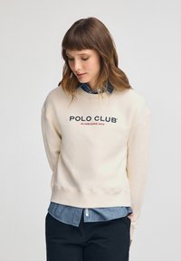 Felpa crema con scollo rotondo e spalle abbassate, con il testo "POLO CLUB" in blu navy e rosso. Indossata sopra una camicia di denim.