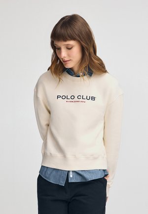 Sudadera crema con cuello redondo y hombros caídos, con el texto "POLO CLUB" en navy y rojo. Superpuesta sobre una camisa de denim.