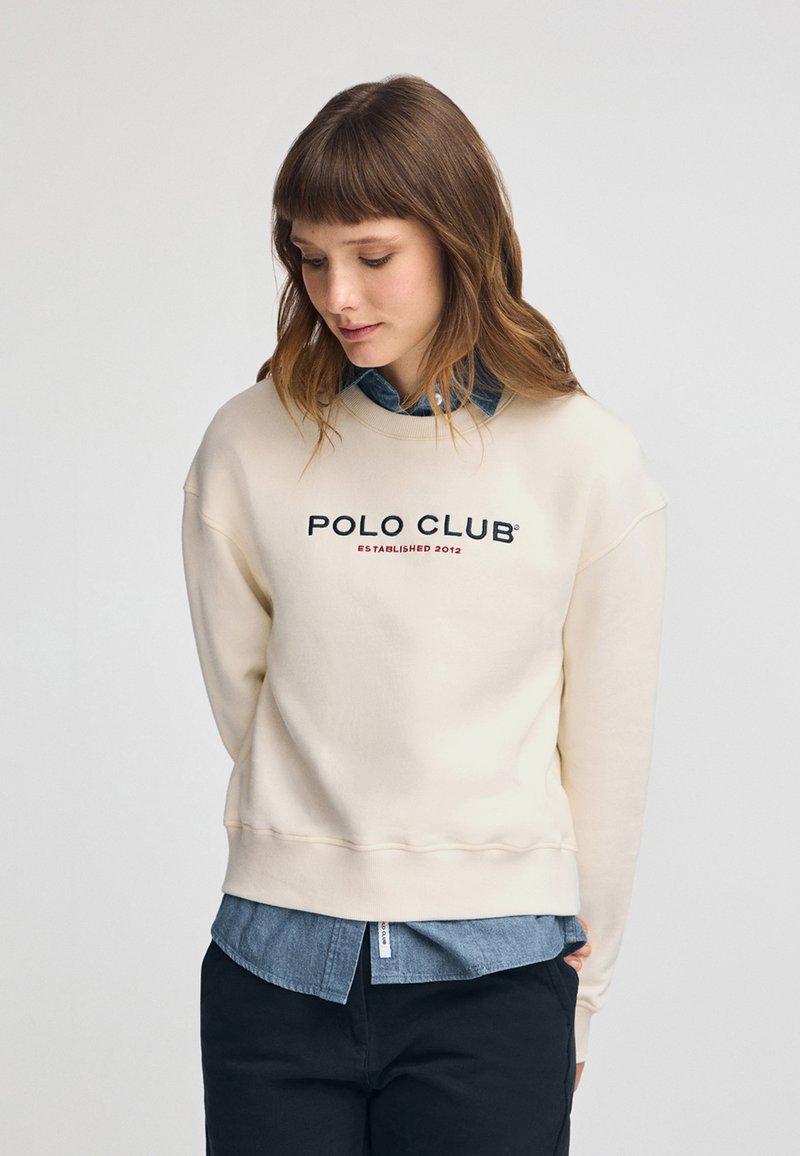 Felpa crema con scollo rotondo e spalle abbassate, con il testo "POLO CLUB" in blu navy e rosso. Indossata sopra una camicia di denim.