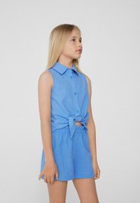 Svetle modrý dvoudílný outfit zahrnující top bez rukávů s knoflíky a uzlem v přední části a odpovídající šortky s elastickým pasem. Hladká textura.