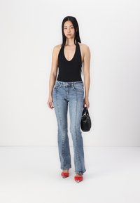 Fekete nyakpántos body, világoskék, harangszárú farmernadrággal, piros magassarkúval és egy kis fekete táskával, fehér háttér előtt.