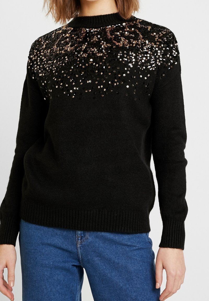 Pull en maille noire avec des sequins brillants dorés, bronze et argentés dispersés sur le haut de la poitrine et les épaules, porté avec un jean bleu.