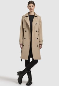 Beige trench coat with a double-breasted design, black buttons, wide lapels, and a belted waist, paired with black leggings and ankle boots.