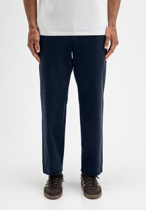 Mann trägt dunkelblaue gerade geschnittene Hose, weißes Hemd und dunkelbraune Sneakers und steht vor einem schlichten weißen Hintergrund.