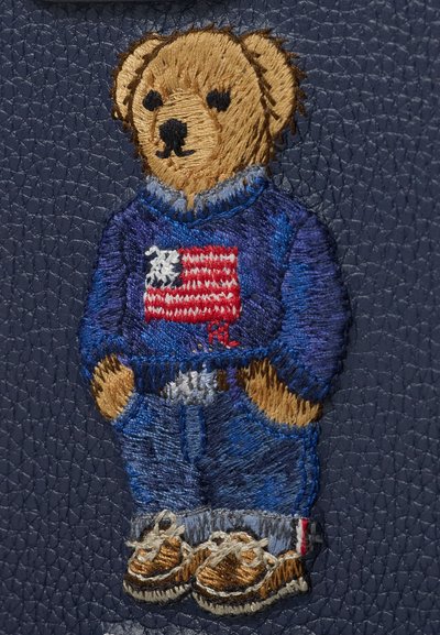 Hímzett mackó kék pulóverben, amerikai zászlóval, kék farmerral és barna cipővel, texturált sötét háttéren.