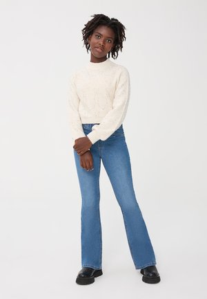 Maglione a maglia crema con motivo texturizzato, abbinato a jeans a zampa d'elefante blu chiaro e stivaletti neri alla caviglia, caratterizzato da una vestibilità comoda e polsini a costine.