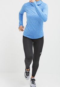 Maglione a maniche lunghe azzurro chiaro con cappuccio; abbinato a leggings neri con logo Nike bianco; scarpe da corsa con accenti neri, bianchi e grigi.