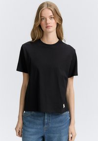 Zwarte katoenen T-shirt met een ronde hals, korte mouwen en een ontspannen pasvorm. Heeft een klein label op de zoom. Draagt met een blauwe spijkerbroek.