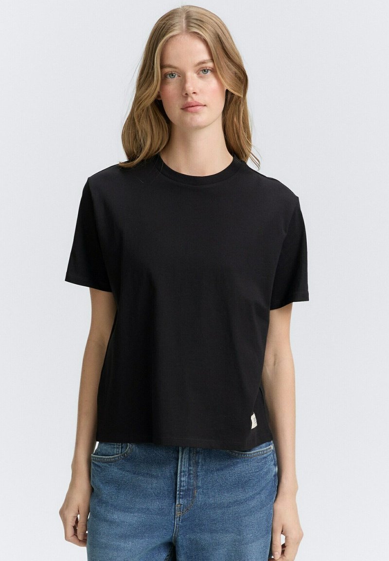 Zwarte katoenen T-shirt met een ronde hals, korte mouwen en een ontspannen pasvorm. Heeft een klein label op de zoom. Draagt met een blauwe spijkerbroek.