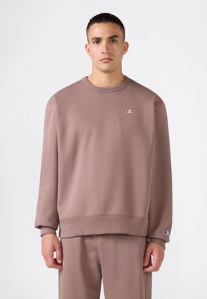 Mann trägt ein hellbraunes Sweatshirt mit langen Ärmeln und passende Hose und steht vor einem weißen, einfarbigen Hintergrund.