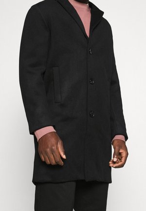 Un modèle porte un manteau noir boutonné par-dessus un pull rose et un pantalon noir, sur un fond blanc uni.