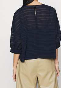 Haut bleu marine avec texture à rayures verticales, manches courtes bouffantes et fermeture dans le dos avec une ouverture en forme de clé. Confectionné dans un matériau léger et transparent.