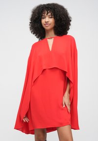 Moteris su garbanotais plaukais, dėvinti ryškiai raudoną, laisvo silueto suknelę su apsiausto formos rankovėmis ir mažais priekyje esančiais raktelio formos iškirptėliais.