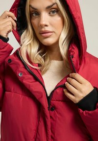 Doudoune rouge avec capuche, dotée d'une fermeture éclair et d'accents noirs aux poignets. Tissu lisse avec une texture matelassée et des boutons-pression.