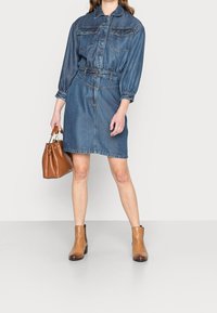 Denim-paita mekko pitkillä hihoilla, kauluksellinen kaula-aukko, vyötärönauha ja kaksi rintataskua. Yhdistetty ruskeisiin nilkkabootseihin ja ruskeaan käsilaukkuun.