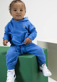 Peuter zittend op een groen blok, gekleed in een blauwe hoodie en broek met witte Nike basketbalschoenen, tegen een effen achtergrond.