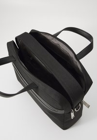 Sac fourre-tout noir avec deux poignées, fermeture éclair et extérieur lisse. L'intérieur comprend un compartiment spacieux et une poche latérale.