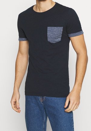 Mann trägt schwarzes Kurzarm-T-Shirt mit meliert grauer Tasche und Ärmelsäumen, kombiniert mit blauen Jeans, gezeigt von Hals bis Hüfte.