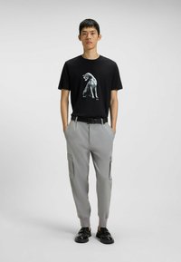 Man in een zwart T-shirt met grafisch ontwerp, lichtgrijze cargobroek met boorden, zwarte riem en zwarte schoenen, staand met zijn handen in zijn zakken.
