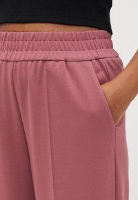 Světlé růžové kalhoty vyrobené z hladkého materiálu, se zpevněným pasem a bočními kapsami. Design zahrnuje jemné sklady v pase.