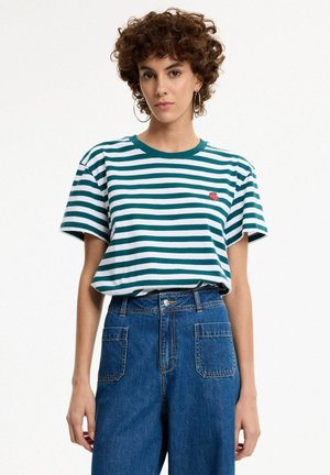 Mujer joven con cabello rizado que lleva una camiseta de rayas verdes y blancas y vaqueros azules de talle alto sobre un fondo blanco.