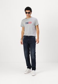Pilkas marškinėliai su "Tommy Jeans" logotipu, tamsiai mėlyni džinsai ir balti sportbačiai. Modelis dėvi juodus akinius nuo saulės. Paprastas, funkcionalus dizainas.