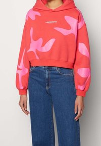 Sudadera corta de color coral con patrones abstractos en rosa. Cuenta con capucha, mangas largas y un logo blanco en el pecho. Se combina con jeans de mezclilla azul oscuro.