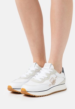 Baskets blanches et grises avec semelles en gomme portées sur des jambes nues, avec un logo marron sur le côté et des lacets blancs.