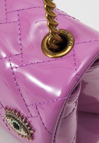 Lila lackläderhandväska med kviltat mönster, guldfärgade detaljer och dekorativt ögonformad utsmyckning på framsidan.