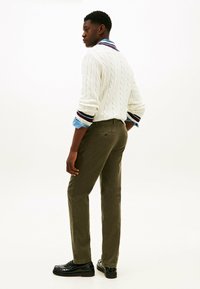 Uomo che indossa un maglione bianco a trecce sopra una camicia blu con le maniche arrotolate, pantaloni verde oliva e mocassini neri, in piedi di profilo.