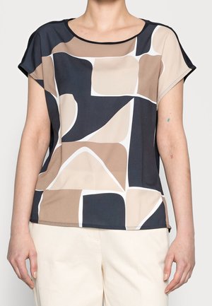 Kobieta ubrana w bluzkę z krótkim rękawem o abstrakcyjnym geometrycznym wzorze w kolorach czarnym, beżowym, brązowym i białym, w połączeniu z kremowymi spodniami.