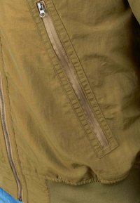 Olijfgroene bomberjack met een gladde textuur, voorzien van een zijvak met rits, ribgebreide manchetten en een elastische tailleband. Minimalistisch ontwerp.