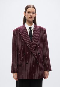 Bordo dvoredni blazer s tankimi navpičnimi črtami, vezenimi logotipi, zarezanimi ovratniki in črnimi gumbi. Stiliziran z belo srajco in kravato.