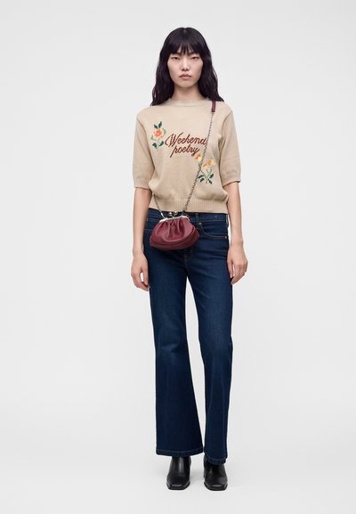 Femme portant un pull beige avec le texte "Weekend poetry", un jean évasé foncé, des bottes noires et un petit sac rouge avec une bandoulière chaîne.