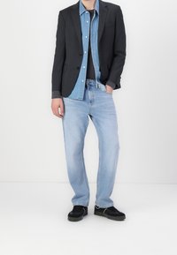 Dunkler Blazer über einem hellen Denim-Hemd, kombiniert mit einem schwarzen T-Shirt und hellblauen Jeans. Schwarze Sneaker runden den Look ab.