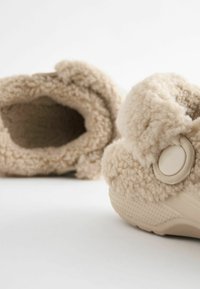 Zapatillas forradas de felpa color beige con forma redondeada, que cuentan con una suave textura esponjosa, un gran botón decorativo y una suela de goma duradera.