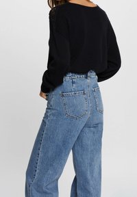Persona vistiendo una blusa negra de manga larga con detalles de encaje en los hombros y unos jeans de mezclilla azul claro de cintura alta, contra un fondo liso.