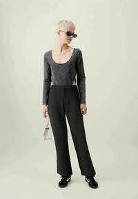 Pessoa com cabelo loiro curto e óculos de sol pretos, vestindo uma blusa cinza de mangas longas, calças pretas e ténis pretos, segurando uma pequena bolsa branca.