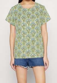 Haut à manches courtes en tissu vert clair avec un motif floral bleu et or. Encolure ronde, coupe décontractée, associé à un short en jean.