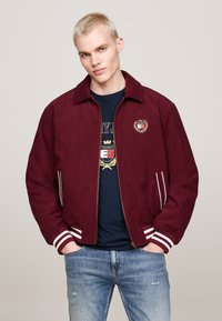 Giacca bomber bordeaux con zip in tessuto morbido, polsini e colletto a righe bianche; logo ricamato sul lato sinistro del petto; indossata sopra una camicia blu.