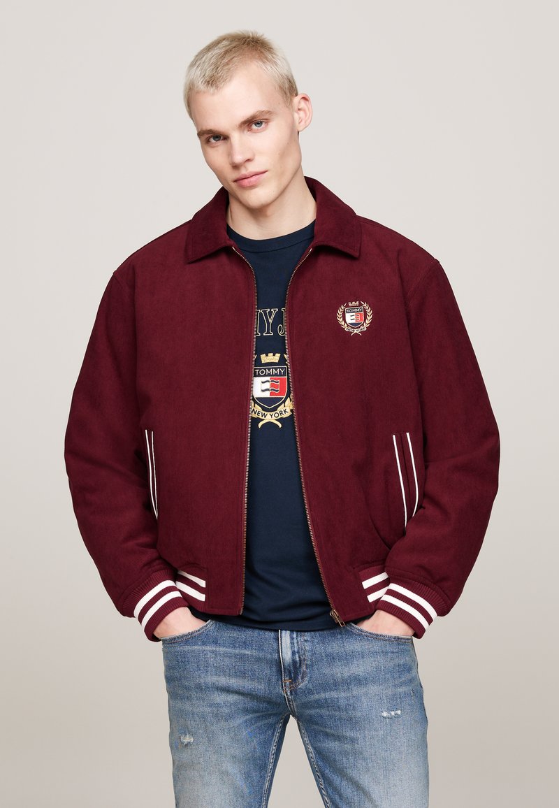 Giacca bomber bordeaux con zip in tessuto morbido, polsini e colletto a righe bianche; logo ricamato sul lato sinistro del petto; indossata sopra una camicia blu.