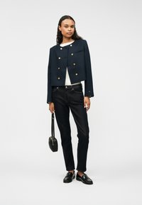 Marineblå kort jakke med gullknapper, hvit topp, mørke jeans og svarte loafers. Har en frontlomme og en liten svart håndveske.